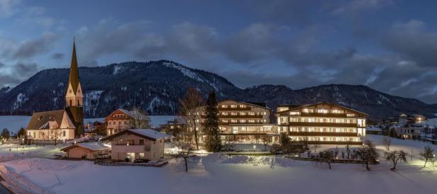 Ein verschneites Dorf mit einer Kirche und einem Hotel vor einer Bergkulisse bei Dämmerung.