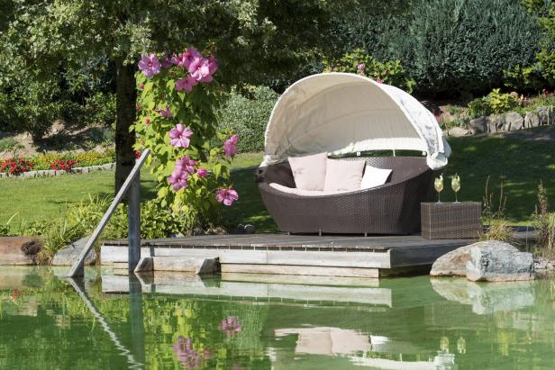 Ein überdachtes Lounge-Bett steht auf einer Holzplattform an einem Teich.