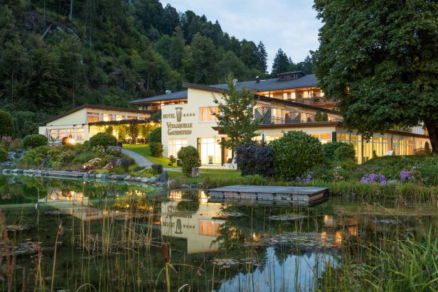 Das Hotel Vitalquelle Gauenstein spiegelt sich im ruhigen Wasser eines Sees.
