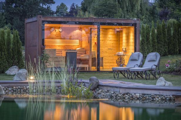 Eine moderne Sauna mit Glasfront steht neben einem Teich und zwei Liegestühlen.
