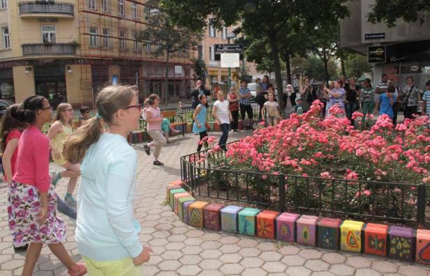Eine Gruppe Kinder läuft auf einem Platz mit einem Rosenbeet und Zuschauern im Hintergrund.