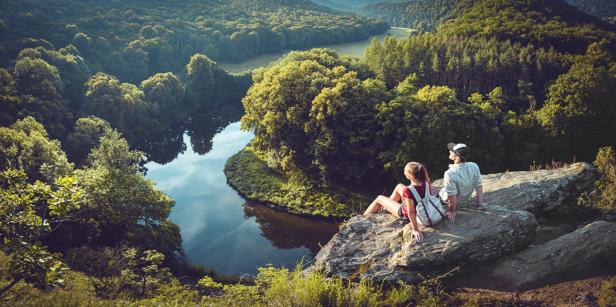 Ein Paar sitzt auf einem Felsen und blickt auf einen Fluss, der sich durch einen grünen Wald schlängelt.