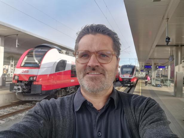 Ein Mann macht ein Selfie vor zwei Zügen der ÖBB an einem Bahnhof.