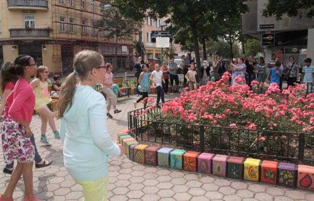 Eine Gruppe von Kindern spielt auf einem Platz mit einem Blumenbeet und bunten Betonblöcken.