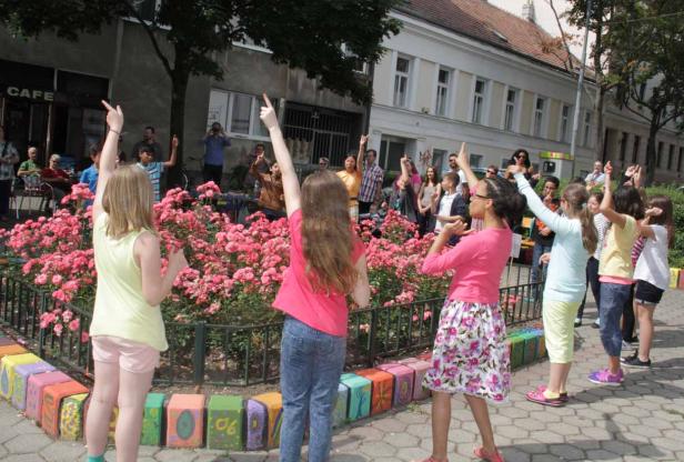 Eine Gruppe Kinder und Erwachsene zeigen mit dem Finger in die Luft vor einem Beet mit rosa Rosen.