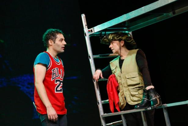 Zwei Schauspieler auf einer Bühne, einer davon auf einer Leiter mit Cowboystiefeln.