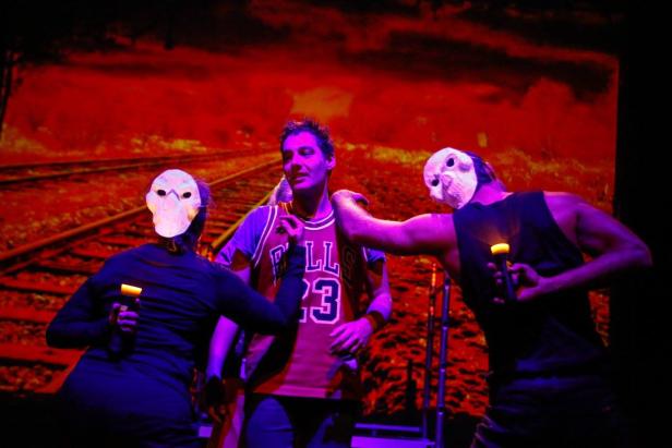 Eine Theaterszene mit einem Mann im Basketballtrikot, umgeben von zwei maskierten Personen vor einem roten Hintergrund.