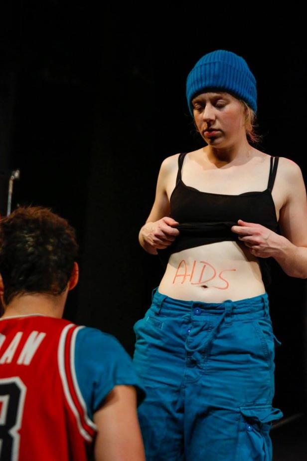 Eine Frau mit blauer Mütze zeigt ihren Bauch, auf dem „AIDS“ geschrieben steht.