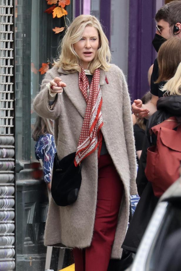 Cate Blanchett in einem beigen Mantel und rotem Schal deutet mit dem Finger.