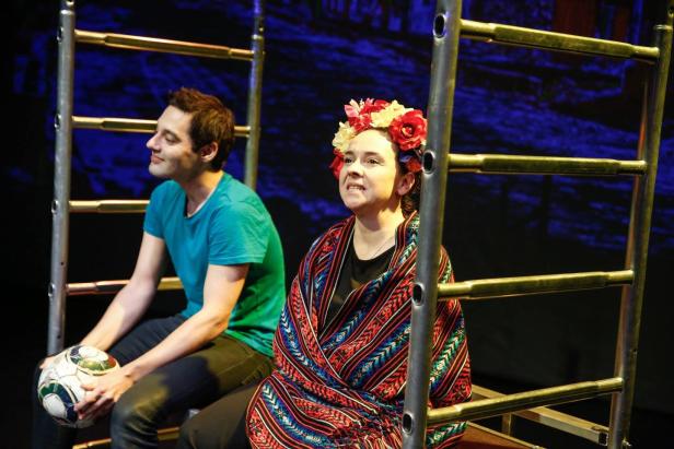 Zwei Schauspieler sitzen auf einer Bühne vor einer Leiter, die Frau mit Blumenkranz und Tuch.