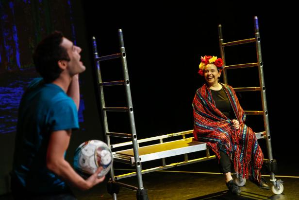 Auf einer Bühne interagieren ein Mann mit einem Fußball und eine Frau mit Blumenkranz auf einer Leiter.