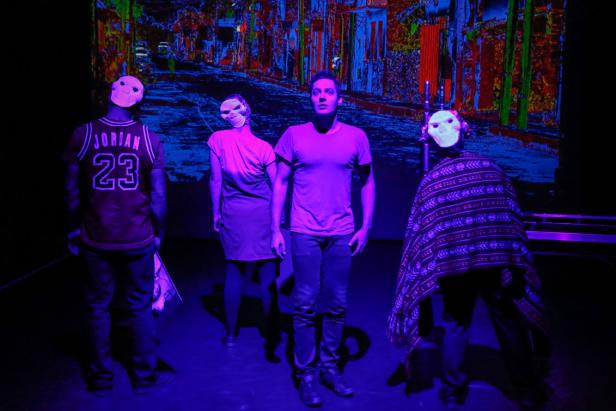 Vier Personen mit Masken stehen vor einer farbenfrohen Projektion einer Stadtansicht.