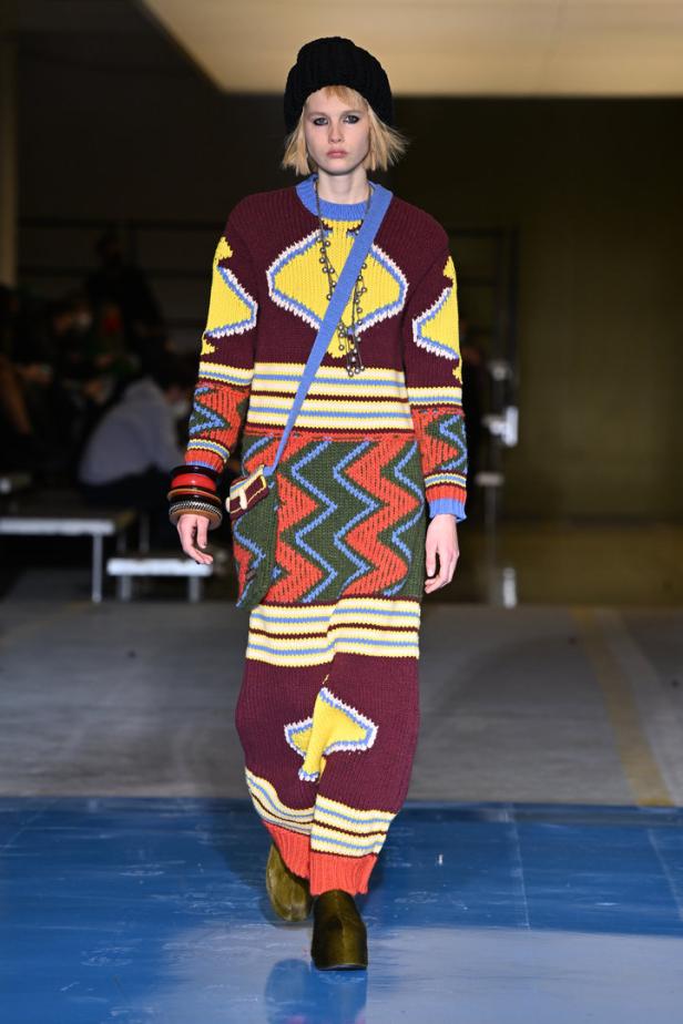 Ein Model präsentiert ein farbenfrohes Strickkleid mit geometrischen Mustern und eine schwarze Mütze auf dem Laufsteg.