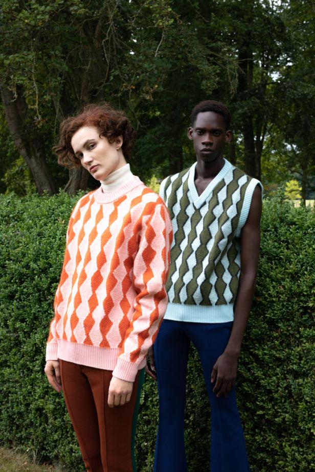 Zwei Models präsentieren farbenfrohe Strickwaren vor einer grünen Hecke.