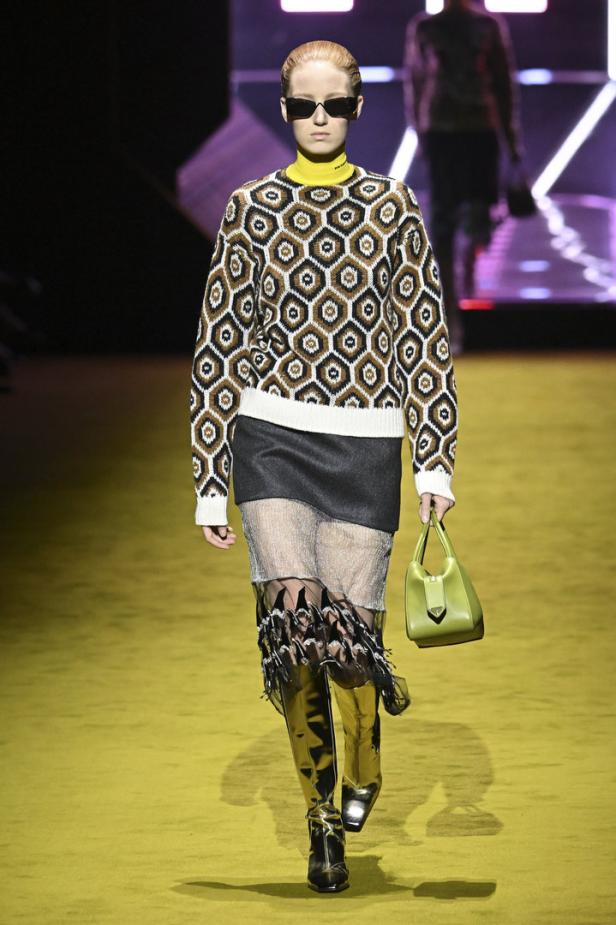 Ein Model auf dem Laufsteg trägt einen gemusterten Pullover, einen Rock und goldene Stiefel.