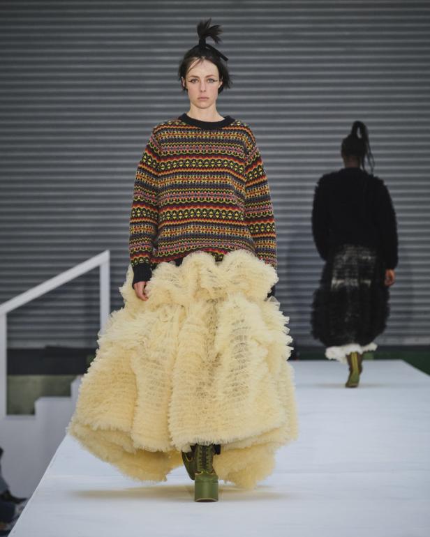 Eine Frau in einem bunten Pullover und einem voluminösen, cremefarbenen Rock auf einem Laufsteg.