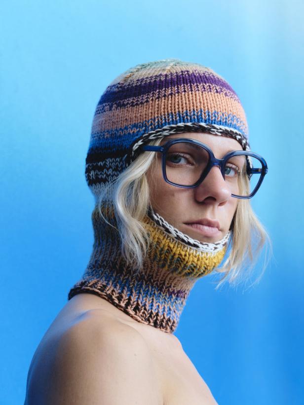 Eine Person mit blonden Haaren trägt eine bunte Sturmhaube und eine blaue Brille vor blauem Hintergrund.