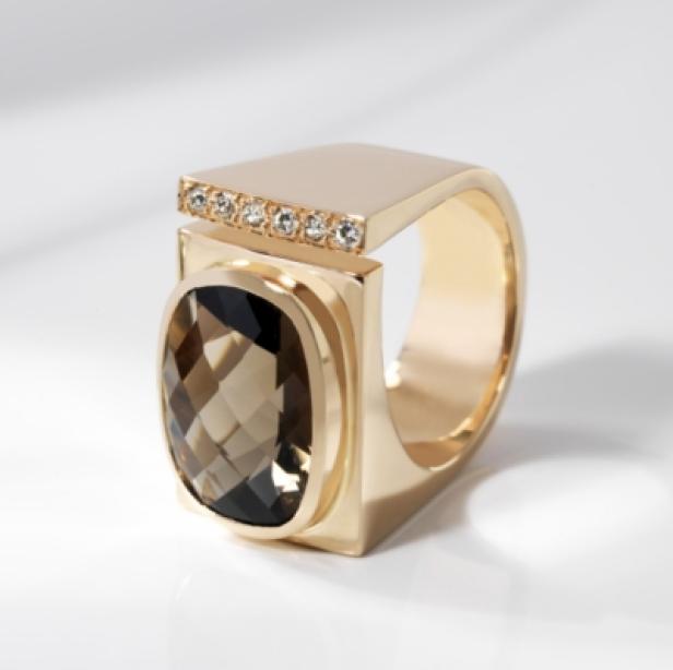 Ein goldener Ring mit einem Rauchquarz und kleinen Diamanten liegt auf einem weißen Untergrund.
