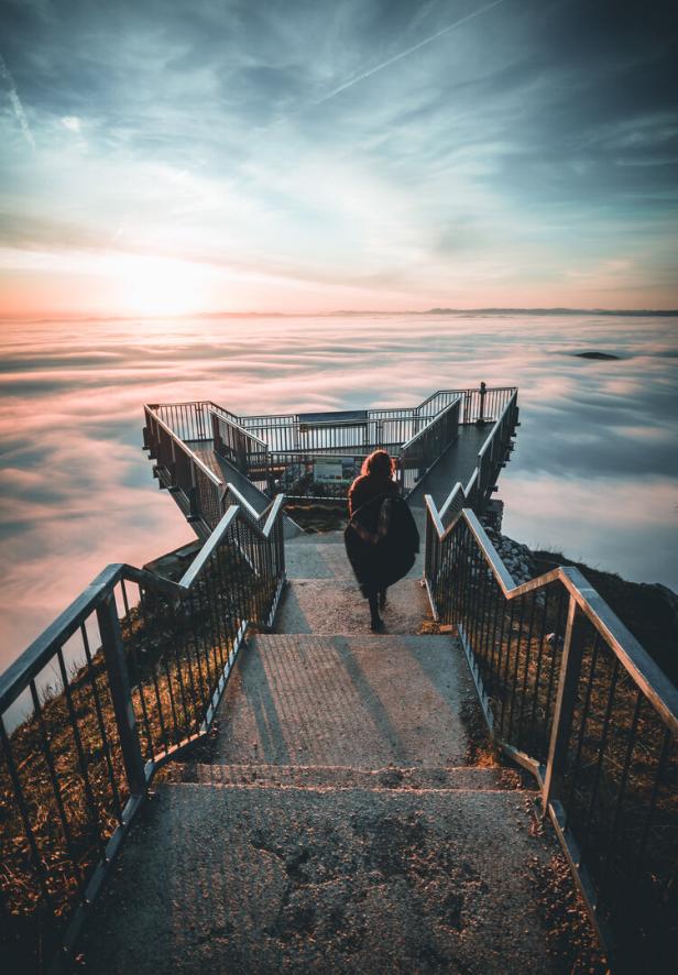 Eine Person geht eine Treppe hinunter zu einer Aussichtsplattform über einer Wolkendecke.