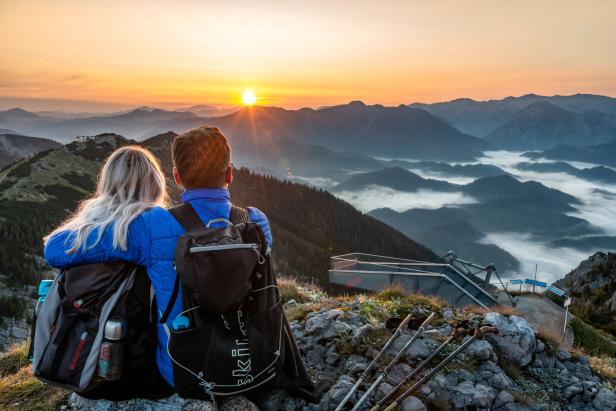 Ein Paar mit Rucksäcken sitzt auf einem Berggipfel und blickt auf den Sonnenaufgang.