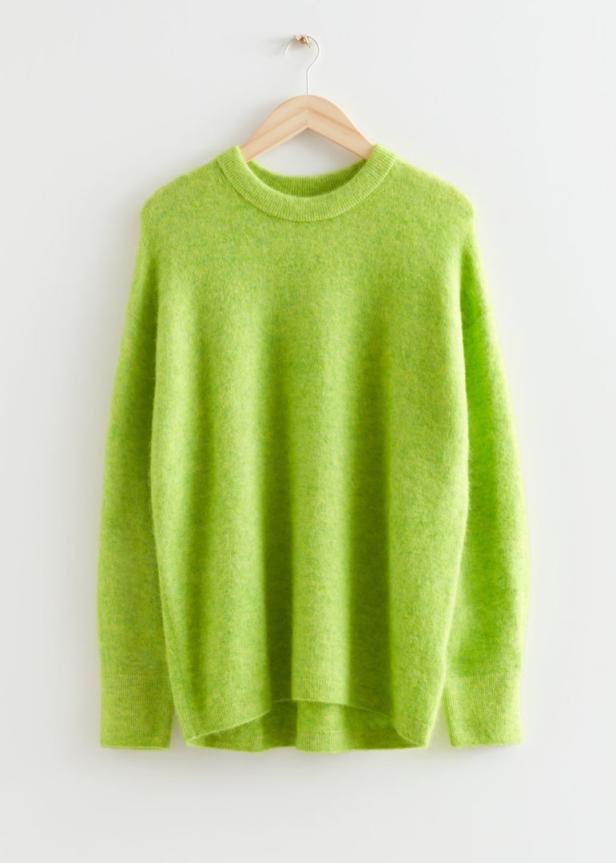 Ein hellgrüner Pullover hängt an einem hölzernen Kleiderbügel vor einer weißen Wand.