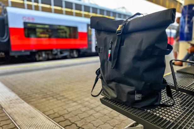 Ein schwarzer Rucksack der Marke „Got Bag“ steht auf einem Bahnsteig vor einem ÖBB-Zug.