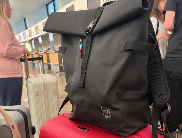 Ein schwarzer Roll-Top-Rucksack steht auf einem roten Koffer am Flughafen.