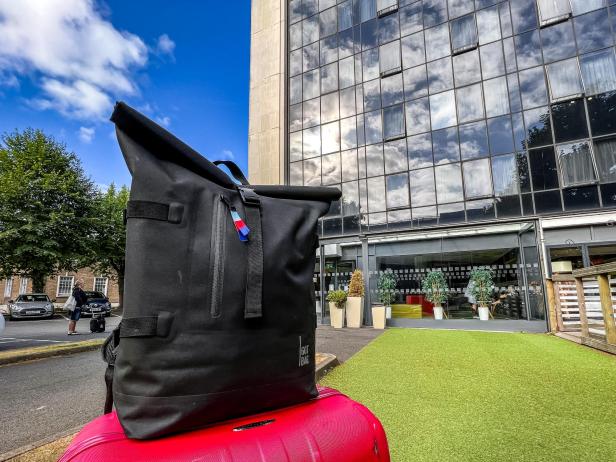 Ein schwarzer Rucksack steht auf einem roten Koffer vor einem modernen Gebäude.