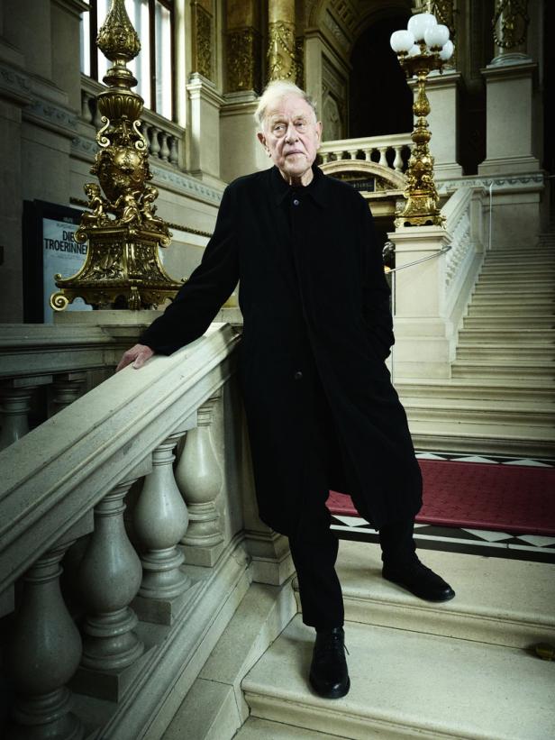 Ein älterer Mann in einem schwarzen Mantel posiert auf einer Treppe in einem opulenten Gebäude.