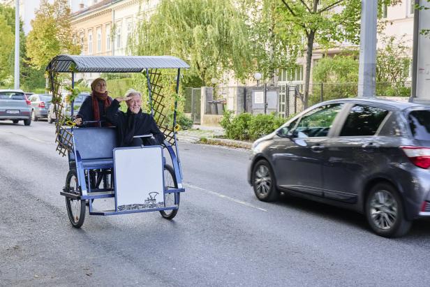 Ein Mann grüßt aus einer Rikscha, während eine Frau sie auf einer Straße entlangfährt.
