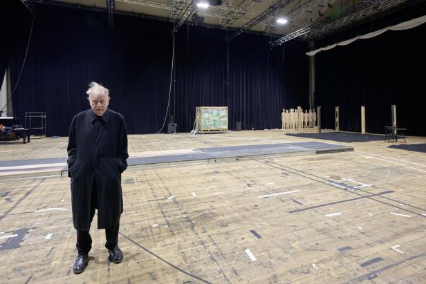 Ein Mann in einem schwarzen Mantel steht in einer großen Halle mit Holzboden.