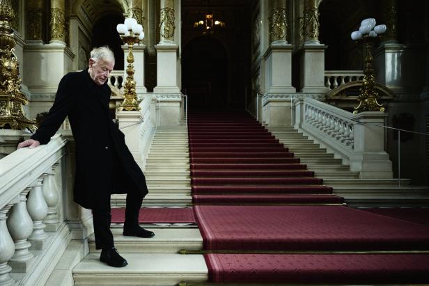 Ein älterer Mann in einem schwarzen Mantel steht auf einer Treppe in einem opulenten Gebäude.