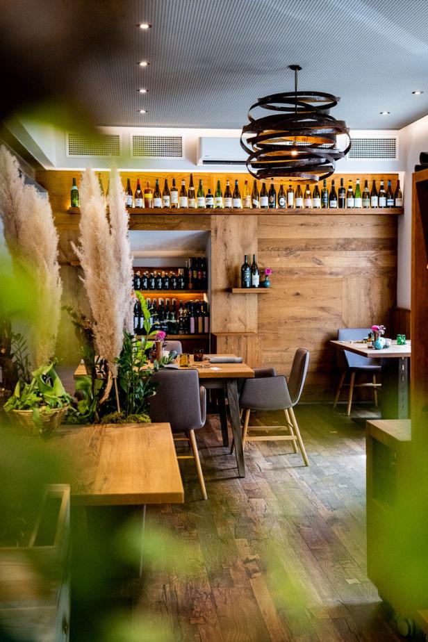 Innenansicht eines Restaurants mit Holztischen, Stühlen und einer Wand mit vielen Weinflaschen.