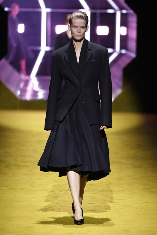 Ein Model in einem schwarzen Blazer und Rock auf einem gelben Laufsteg.