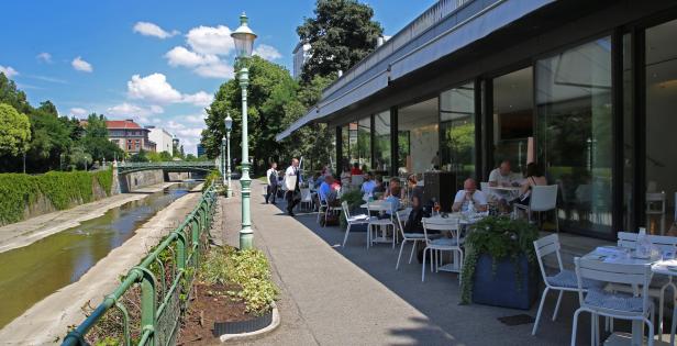 Ein Restaurant am Flussufer mit Gästen im Freien an einem sonnigen Tag.