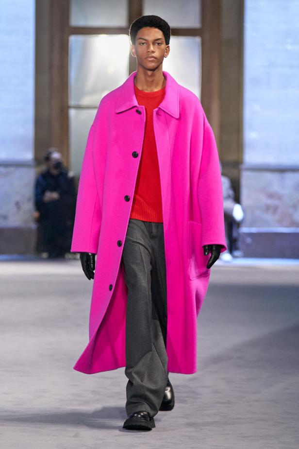 Ein Mann in einem pinkfarbenen Mantel, rotem Pullover und grauer Hose läuft auf einem Laufsteg.