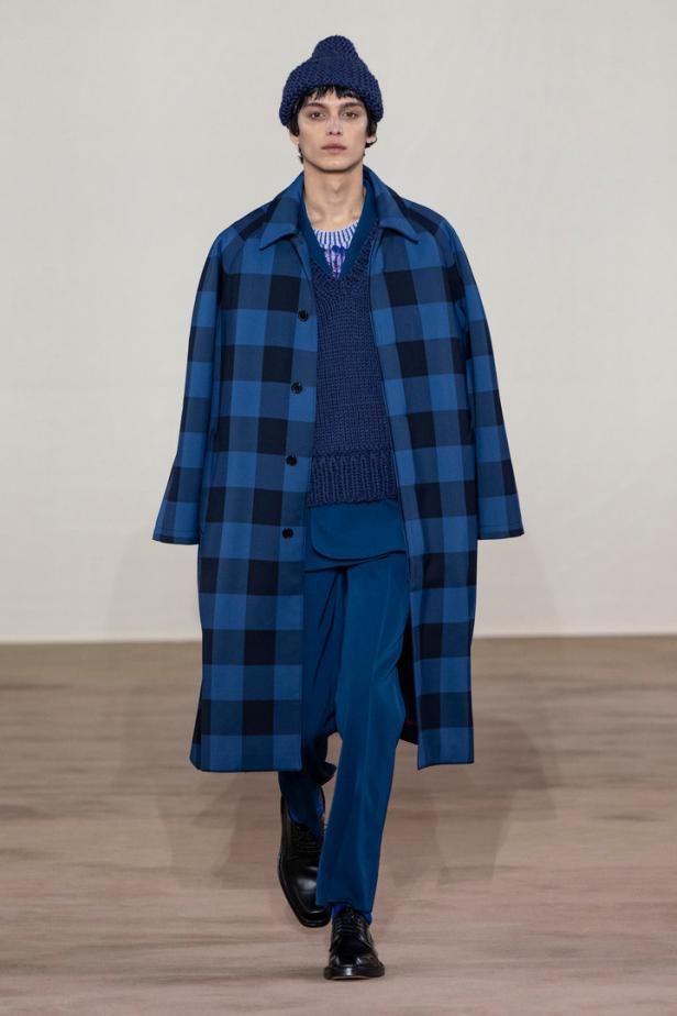 Ein Mann in blauem Outfit mit kariertem Mantel und Strickmütze auf dem Laufsteg.