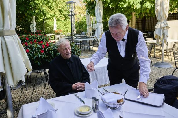 Ein älterer Mann sitzt an einem Tisch im Freien, während ein Kellner den Tisch deckt.