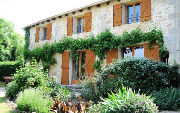 Ein zweistöckiges Steinhaus mit braunen Fensterläden und üppigem Grün im Garten.