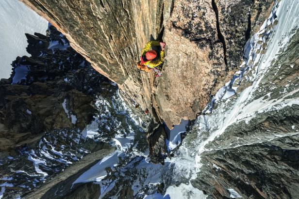 Ein Bergsteiger klettert eine steile Felswand in einer verschneiten Berglandschaft hinauf.