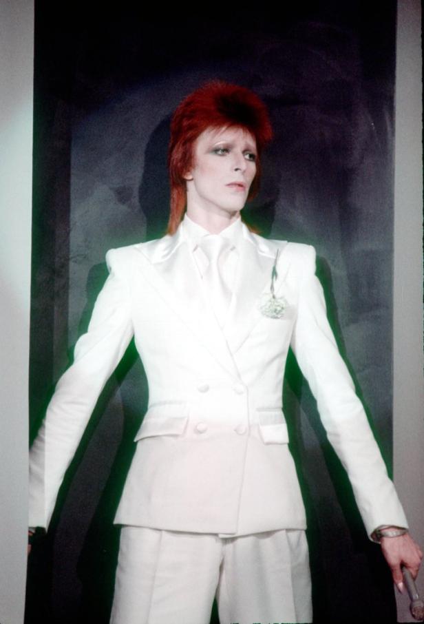 David Bowie in einem weißen Anzug mit rotem Haar und einem Blumenanstecker.