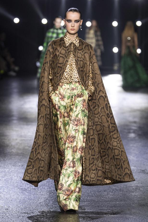 Eine Frau in einem Schlangenmuster-Cape, Leopardenbluse und Rosenhose auf einem Laufsteg.