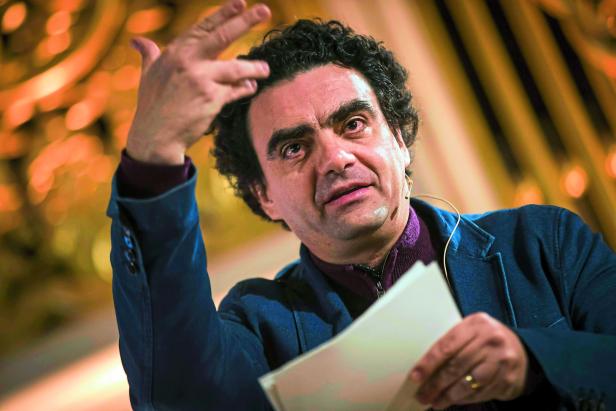 Ein Mann mit lockigem Haar gestikuliert während eines Vortrags mit einem Blatt Papier in der Hand.