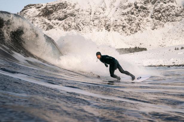 Ein Surfer reitet eine Welle vor einer schneebedeckten Bergkulisse.