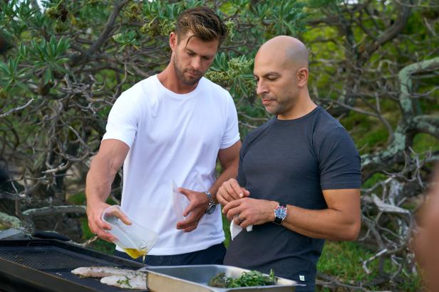 Chris Hemsworth gießt Öl über Fisch auf einem Grill, während ein anderer Mann zusieht.