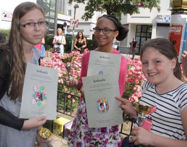 Drei Mädchen präsentieren Urkunden und kleine goldene Trophäen vor einem Blumenbeet.
