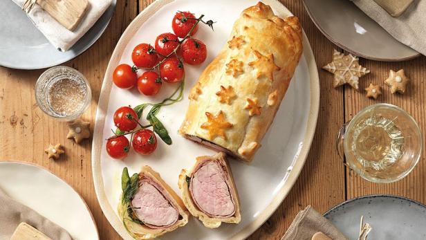 Ein Schweinefilet im Teigmantel mit Salbei und Tomaten auf einem festlich gedeckten Tisch.