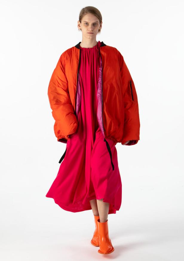 Eine Frau trägt eine orangefarbene Bomberjacke über einem pinkfarbenen Kleid und orangefarbene Stiefel.