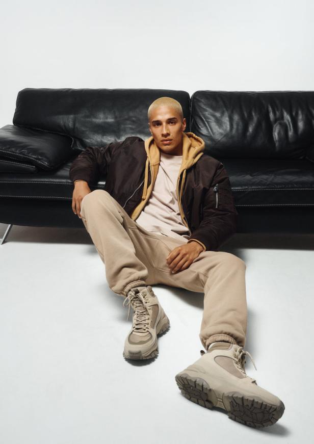 Ein Mann sitzt vor einem schwarzen Ledersofa, bekleidet mit Jacke, Hoodie, Jogginghose und Stiefeln.