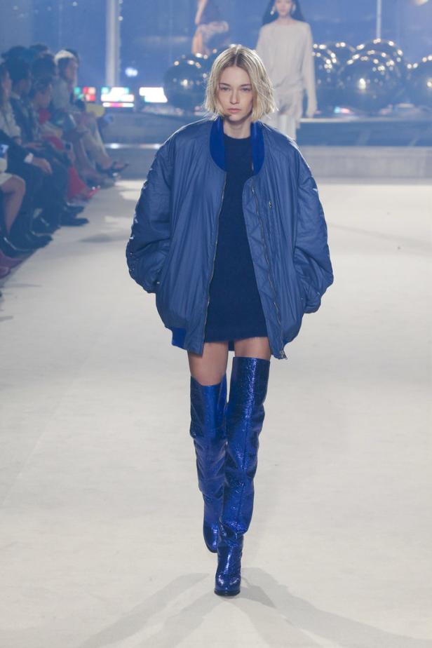 Eine Frau mit blonder Bob-Frisur läuft in einem blauen Bomber und glänzenden Stiefeln über den Laufsteg.